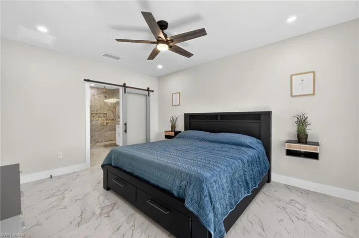 Bedroom with a barn door, ceiling fan, ensuite bath, recessed lighting, and light marble finish flooring
