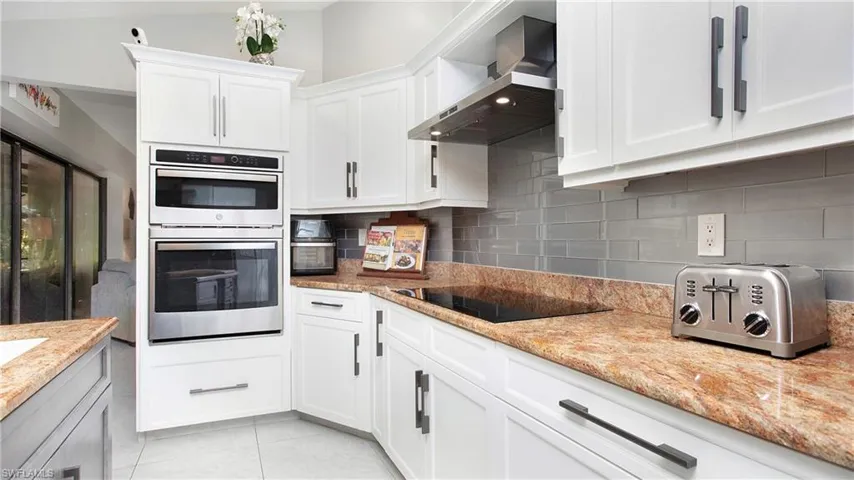 Kitchen featuring wall chimney exhaust hood, double oven, black electric stovetop, white cabinetry, and backsplash