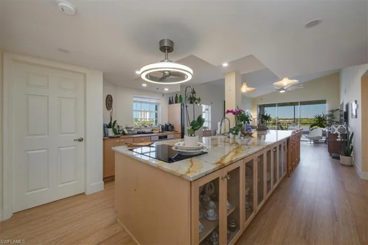 Kitchen with light stone counters, light wood-type flooring, glass insert cabinets, a ceiling fan, and a center island with sink