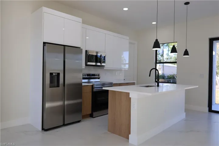 Dual tone kitchen featuring stainless steel appliances, an island with sink, two tone cabinets, light marble finish floors, and pendant lighting