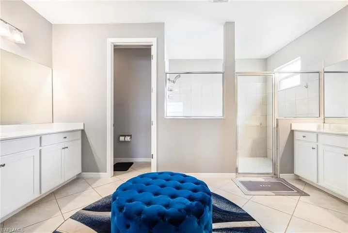 Bathroom featuring dual vanity areas with white cabinetry and light countertops, a glass-enclosed shower with tiled walls, and light-toned tile flooring