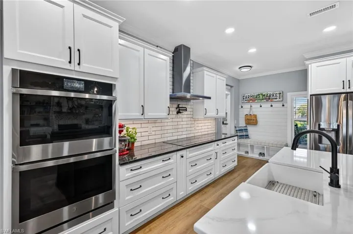 Kitchen with stainless steel appliances, white cabinetry, dark stone countertops, wall chimney range hood, and crown molding