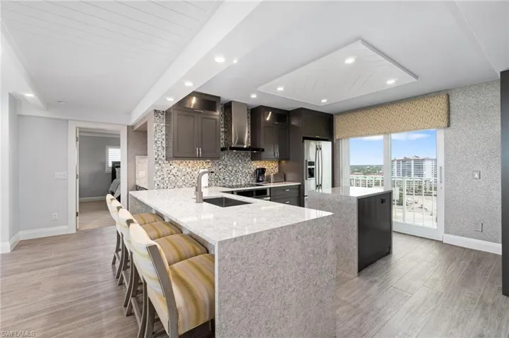Kitchen featuring a peninsula, light stone counters, wall chimney exhaust hood, stainless steel refrigerator with ice dispenser, and a breakfast bar area