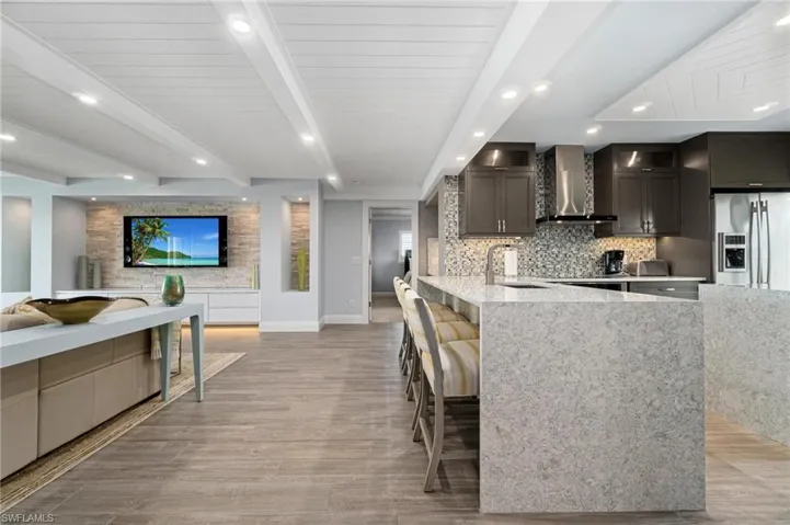 Kitchen with beam ceiling, stainless steel fridge with ice dispenser, a kitchen bar, wall chimney range hood, and light stone counters