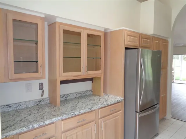 Kitchen featuring freestanding refrigerator, light stone counters, glass insert cabinets, and light brown cabinets