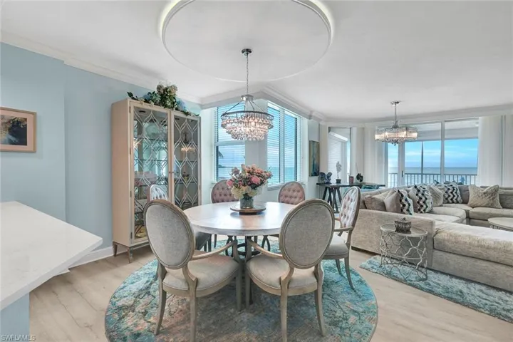 Open-concept area featuring light-tone wood-finish flooring, recessed circular ceiling detail, dual crystal chandeliers, floor-to-ceiling windows, and sliding glass door access to a balcony