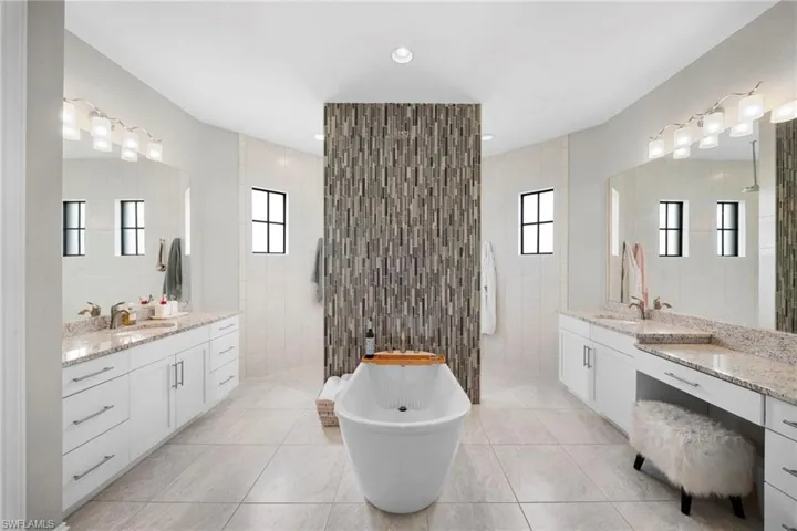 Freestanding soaking tub with a tiled accent wall, dual vanity areas with granite countertops, and light-toned floor tiling