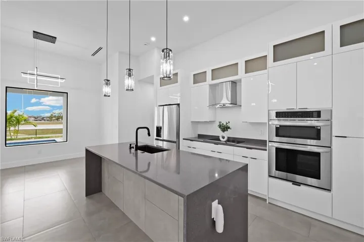 Kitchen featuring wall chimney range hood, stainless steel appliances, a center island with sink, hanging light fixtures, and sink