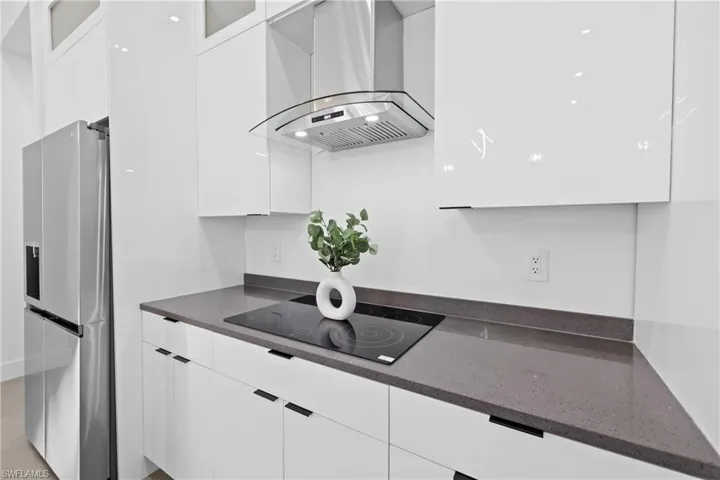 Kitchen featuring dark stone counters, black electric stovetop, stainless steel fridge, wall chimney exhaust hood, and white cabinets