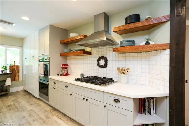 Kitchen with open shelves, wall chimney range hood, light countertops, light wood-style floors, and backsplash