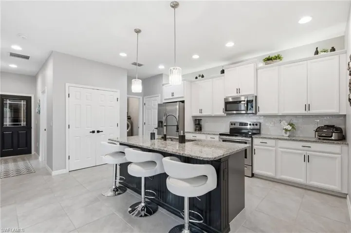 Modern kitchen featuring a granite-topped island with pendant lighting, stainless steel appliances, white cabinetry with black hardware, a tiled backsplash, and light-toned tile flooring