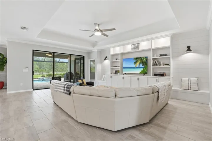 Spacious common area featuring light-toned tile flooring, tray ceiling with a ceiling fan, and built-in shelving with cabinetry