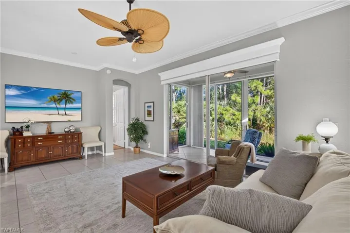 Neutral-toned living space featuring large-format ceramic tile flooring, crown molding, a multi-blade ceiling fan, and sliding glass doors with custom valance