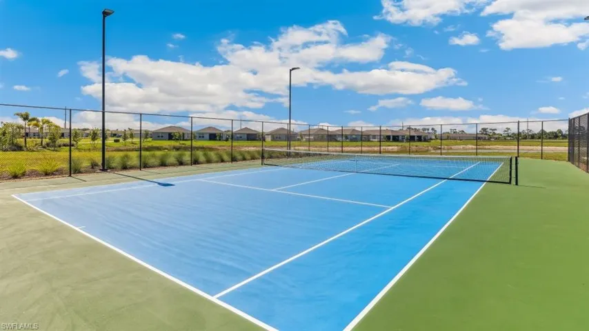 Tennis court featuring a blue playing surface with white lines and green surrounds, enclosed by a black chain-link fence