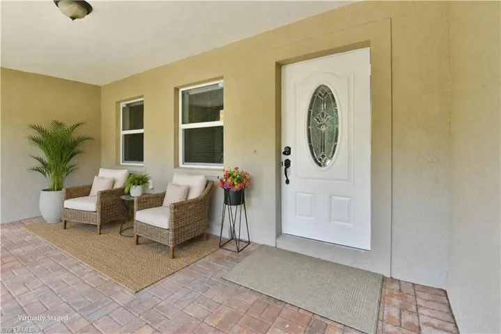 Covered porch with paver flooring, stucco exterior, white entry door with decorative glass insert, and two windows