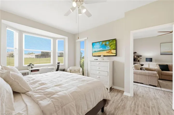 Spacious bedroom featuring light wood-finish flooring, a ceiling fan with integrated lighting, and a wide bay window with horizontal blinds offering exterior views