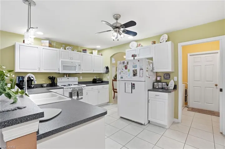 Kitchen featuring arched walkways, dark countertops, white cabinets, white appliances, and ceiling fan