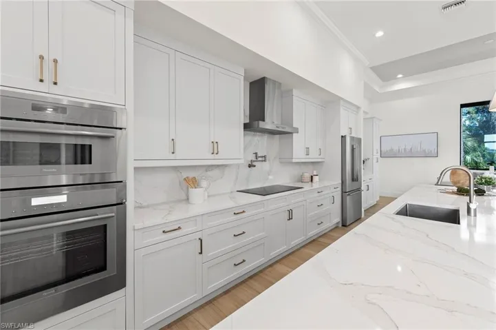Kitchen featuring wall chimney range hood, sink, backsplash, stainless steel appliances, and white cabinets