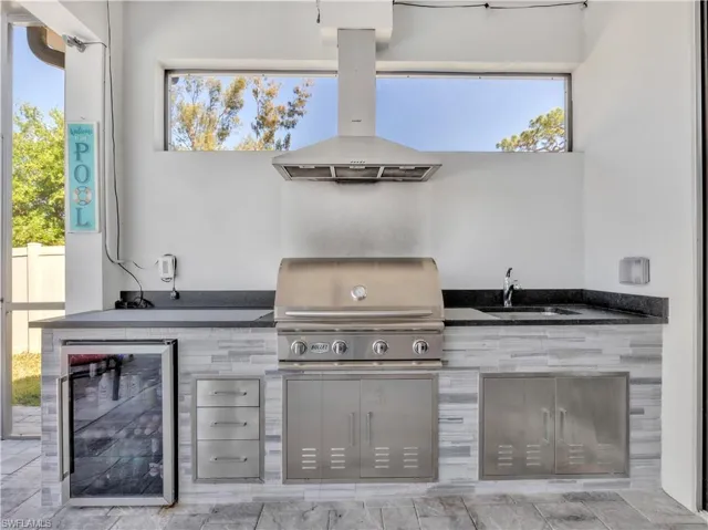 Outdoor kitchen featuring a built-in stainless steel grill, integrated sink, beverage refrigerator, and dark countertop surfaces