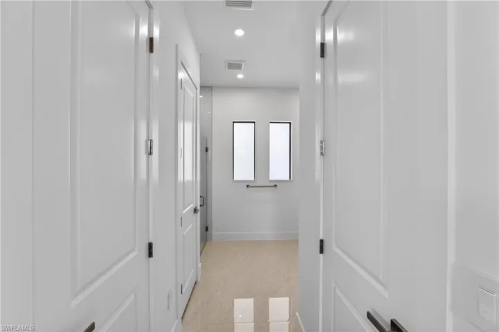 Bright hallway featuring recessed lighting, light-toned tile flooring, white paneled doors with dark hardware, and two vertical windows at the far end