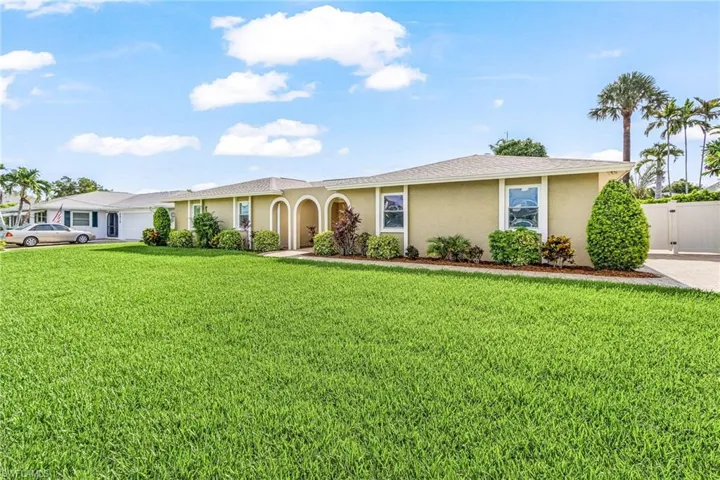 Spacious front lawn with mature landscaping, stucco exterior, arched entryways, white-framed windows, and a privacy fence
