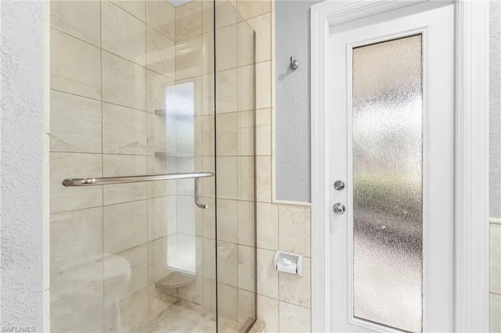 Glass-enclosed shower featuring light-toned square tile, a built-in corner shelf, and a brushed metal handle