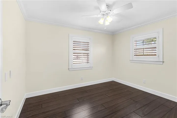 Room featuring dark wood-finish flooring, two windows with white plantation shutters, crown molding, white baseboards, and a white ceiling fan with integrated lighting