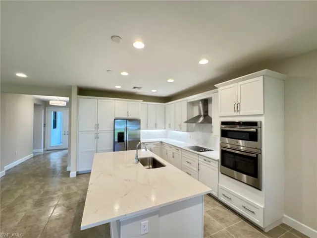 Kitchen with white cabinetry, appliances with stainless steel finishes, recessed lighting, wall chimney exhaust hood, and light stone counters