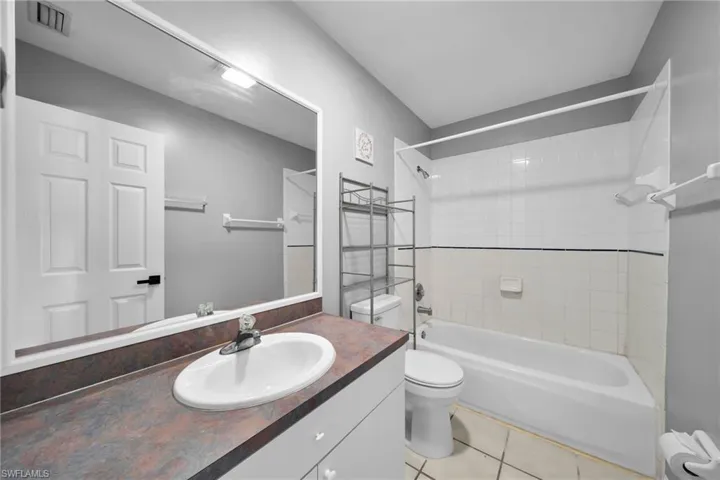 Bathroom featuring a vanity with an oval sink and a dark-toned countertop, a full-width wall mirror, a white paneled door with black hardware, a shower-tub combination with white tile surround, and light-toned floor tiles
