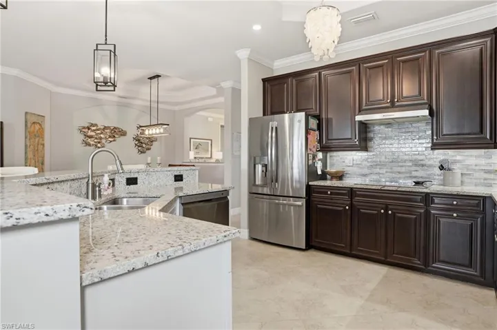 Kitchen featuring crown molding, suspended lighting, stainless steel appliances, light stone counters, and dark wood finish cabinetry