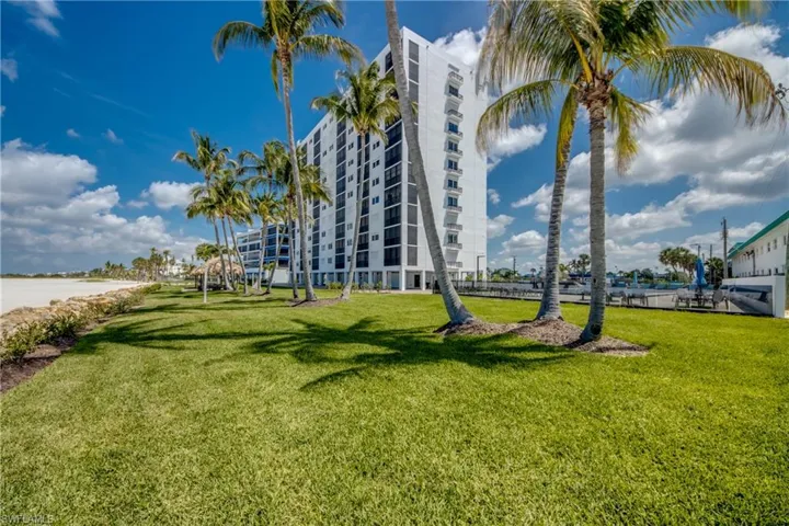 Waterfront property featuring a multi-story building, lush green lawn, mature palm trees, sandy beach access, and a boat dock