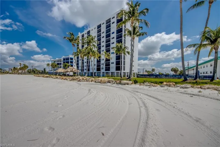 Expansive sandy beach with a high-rise building backdrop, featuring numerous palm trees, a thatched-roof structure, and a manicured lawn area