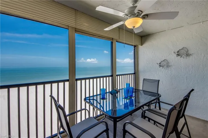 Expansive balcony featuring a white textured wall, ceiling fan with integrated lighting, full-height windows, and a black railing overlooking the ocean