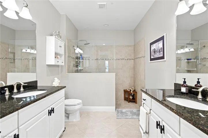 Spacious bathroom featuring dual vanities with dark granite countertops and undermount sinks, a walk-in shower with glass enclosure and tiled walls, light-toned floor tiling, and white cabinetry