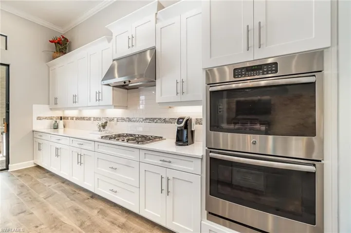 Kitchen featuring white cabinets, crown molding, decorative backsplash, light wood-type flooring, and appliances with stainless steel finishes