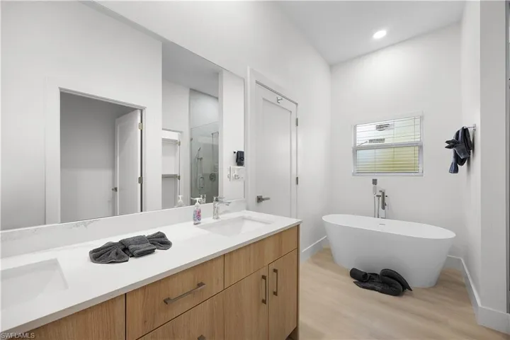 The bathroom features a double vanity with light wood cabinetry and a white countertop, a freestanding white bathtub with a floor-mounted faucet, and light wood-tone flooring