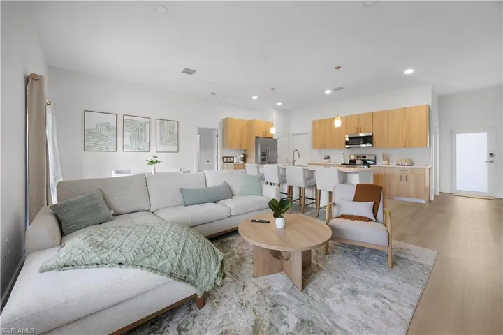 Open concept living area featuring light-toned flooring, recessed lighting, and a kitchen with light wood cabinetry, stainless steel appliances, and a breakfast bar with seating