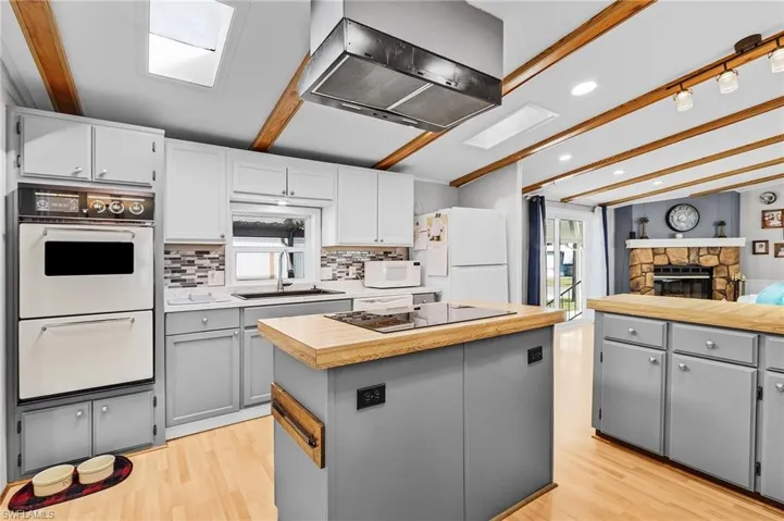 Kitchen featuring a center island, wall chimney exhaust hood, gray cabinetry, beam ceiling, and white appliances