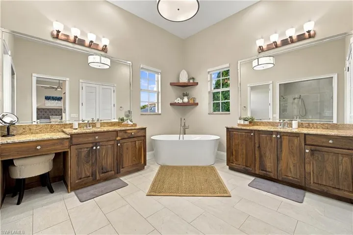 Freestanding soaking tub, dual wood-finish vanity cabinetry, stone-surface countertops, large wall-mounted mirrors, and tiled flooring