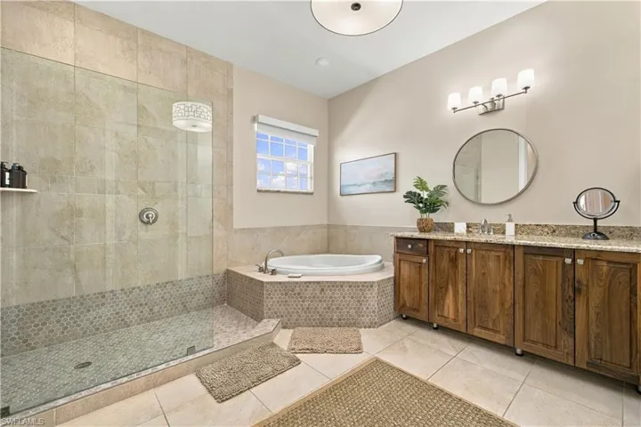 Walk-in shower with stone tile surround, soaking tub with mosaic tile skirt, wood-finish cabinetry, stone countertop, and circular vanity mirror