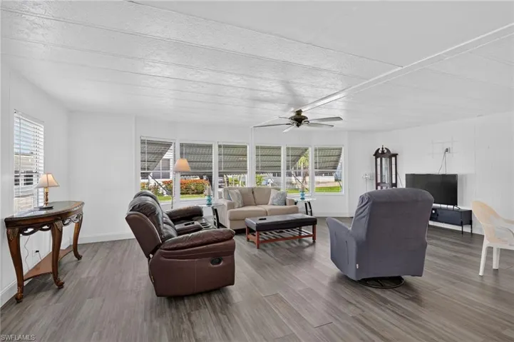 Spacious living area featuring wood-finish flooring, white paneling, multiple windows with blinds, and a ceiling fan with light fixture