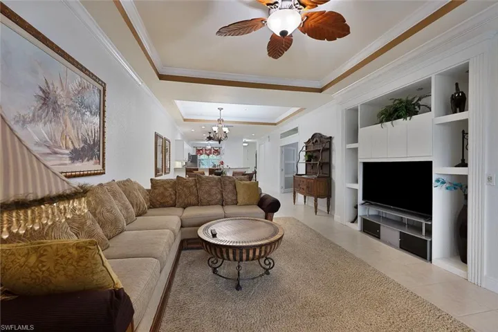 Tiled living room with ornamental molding, a tray ceiling, a chandelier, a ceiling fan, and built in features