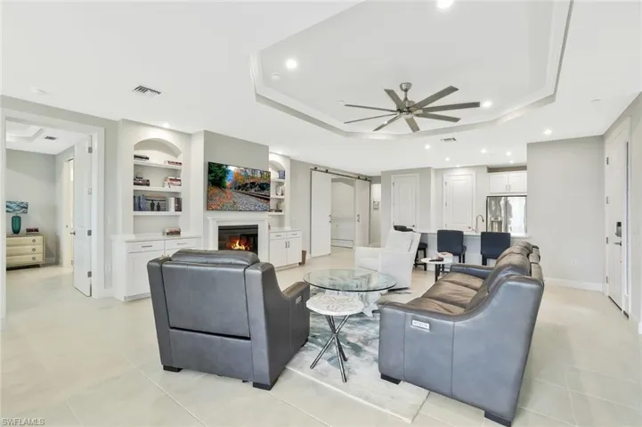 Living room featuring built in shelves, a glass covered fireplace, ceiling fan, a tray ceiling, and recessed lighting