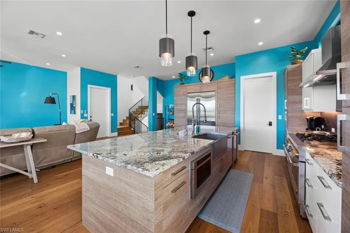 Kitchen with an island with sink, built in appliances, pendant lighting, wood flooring, and wall chimney range hood