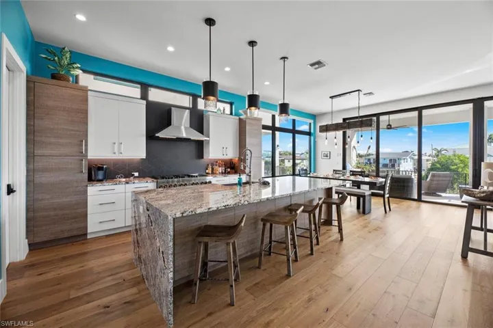 Kitchen with wall chimney range hood, hanging light fixtures, a center island with sink, tasteful backsplash, and white cabinetry