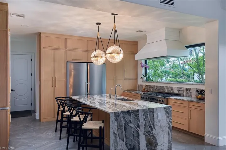 Gourmet kitchen featuring a large island with waterfall countertop, stainless steel appliances, a wide window overlooking tropical foliage, custom wood cabinetry, and sleek pendant lighting