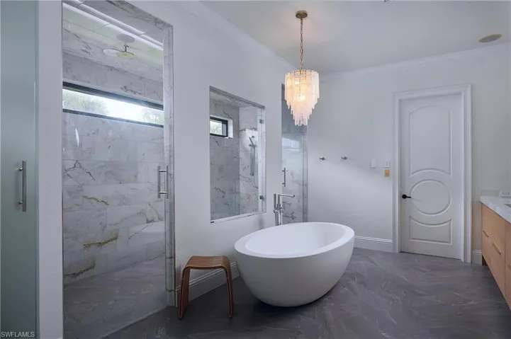 Spa-inspired bathroom featuring a freestanding oval soaking tub, a spacious glass-enclosed shower with marble-look tile, dark stone-look flooring, a contemporary chandelier, and a white oak custom vanity with a stone countertop