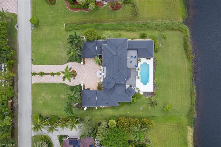 Estate property featuring a paver driveway with a circular turnaround, lush green lawns, mature palm trees, a dark tile roof, and a private swimming pool with an expansive deck bordering a body of water