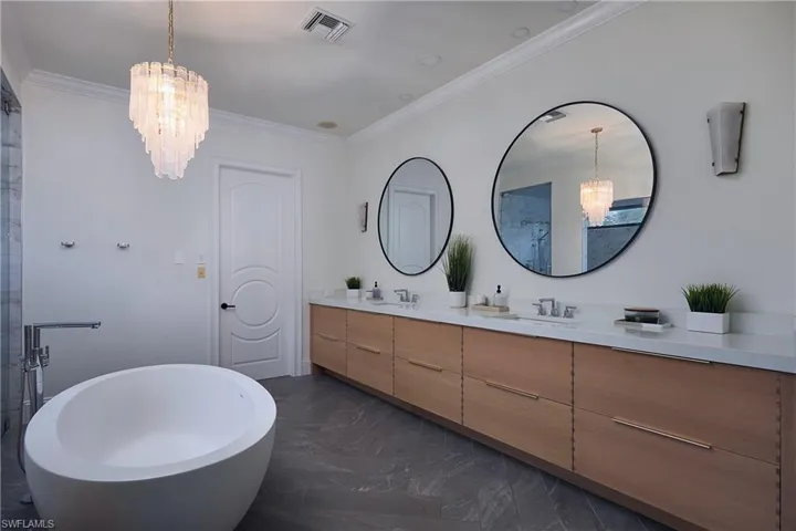 Contemporary bathroom featuring a freestanding soaking tub, a dual vanity with a light wood-finish cabinetry, round framed mirrors, and a striking chandelier