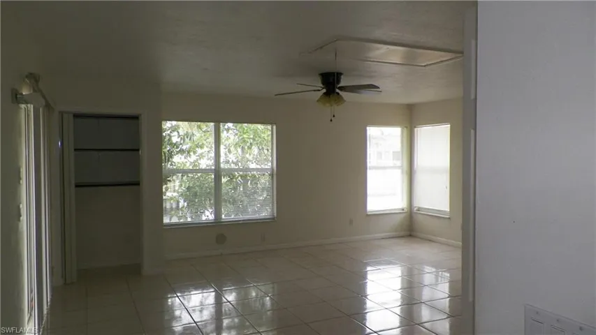Spacious room featuring light-toned square tile flooring, multiple windows providing ample natural light, a ceiling fan with integrated lighting, and a built-in closet with shelving
